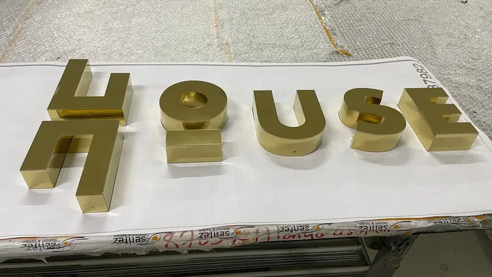 Gold mirror stainless steel letters spelling HOUSE laid out in the workshop